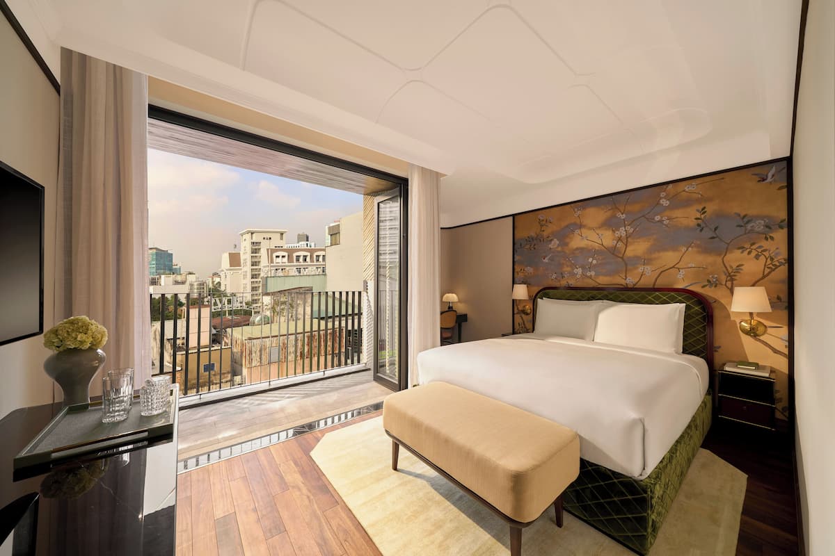 Une chambre d’hôtel avec un grand lit, un bureau agrémenté d’un vase, et une vue sur la ville depuis un balcon.