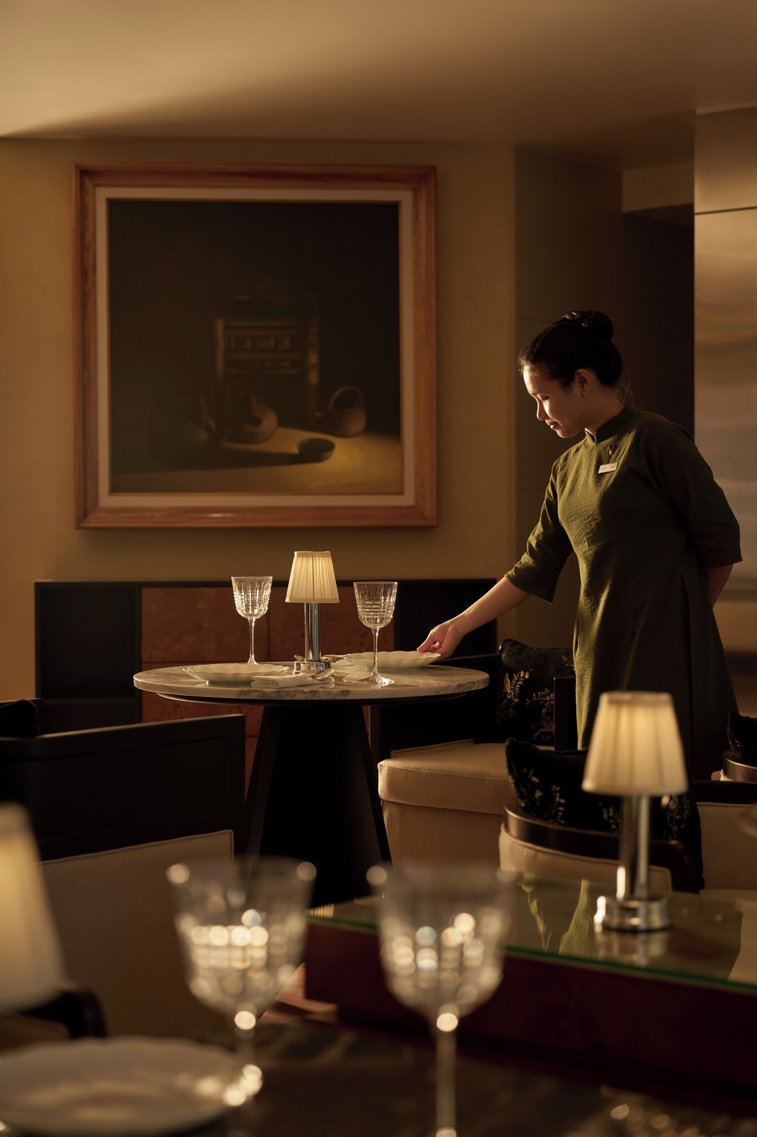 A dining area with a table set for two, a painting on the wall, and a person standing by the table.