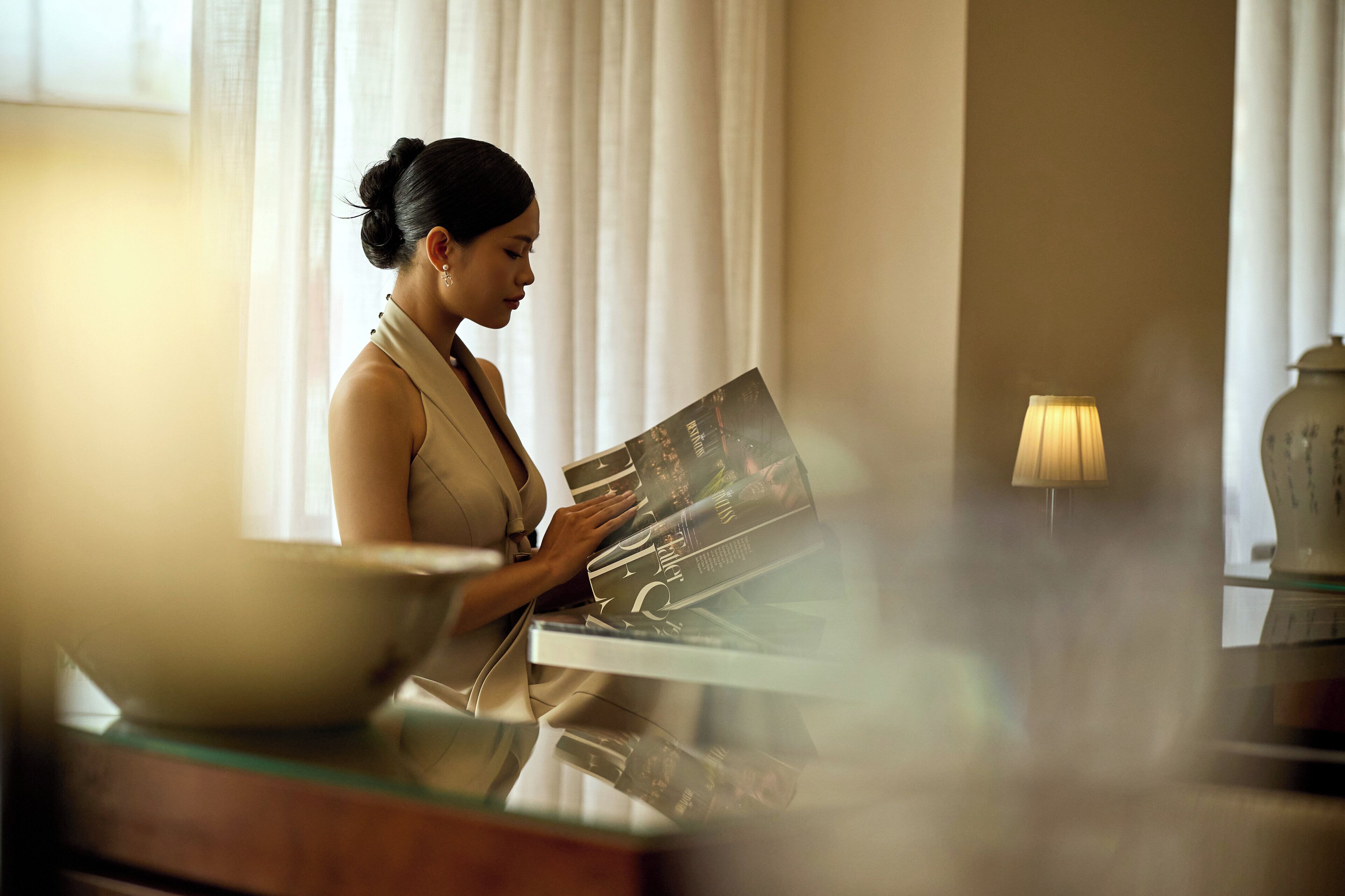 Una persona leyendo una revista en una mesa, con un bol y una lámpara, en una habitación con cortinas y un florero.