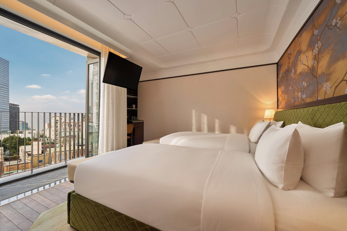 A hotel room with a large bed, a green headboard, a city view, and a wall-mounted TV.