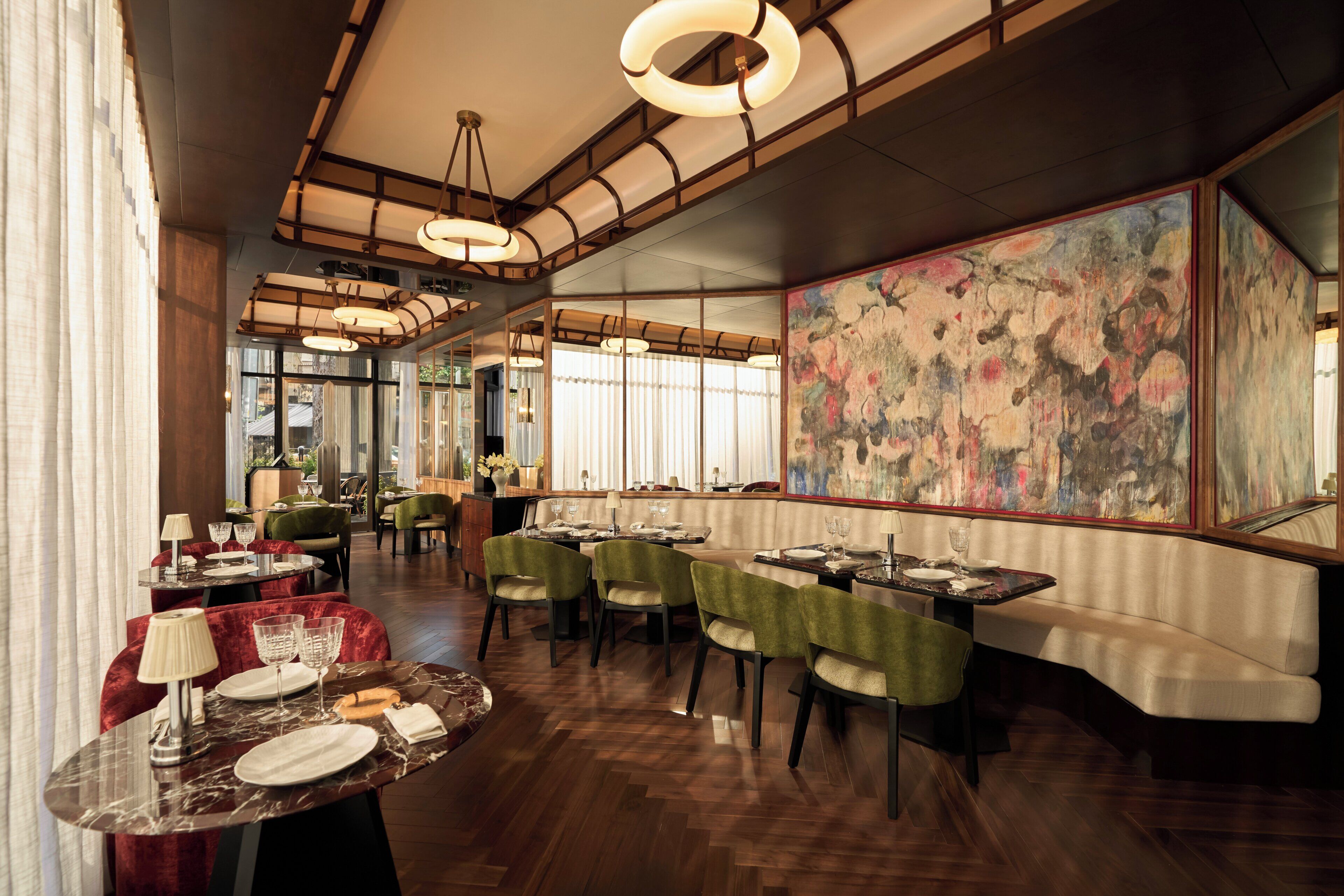 A modern restaurant interior with wooden flooring, green upholstered chairs, marble tables, and a large floral painting on the wall.
