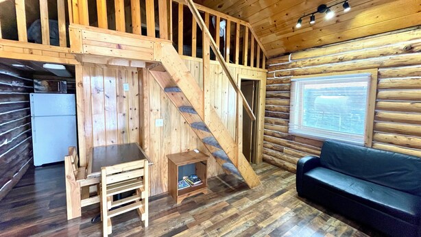 A cabin interior with a wooden staircase, a small kitchen area, a seating area with a couch, and a window with blinds.
