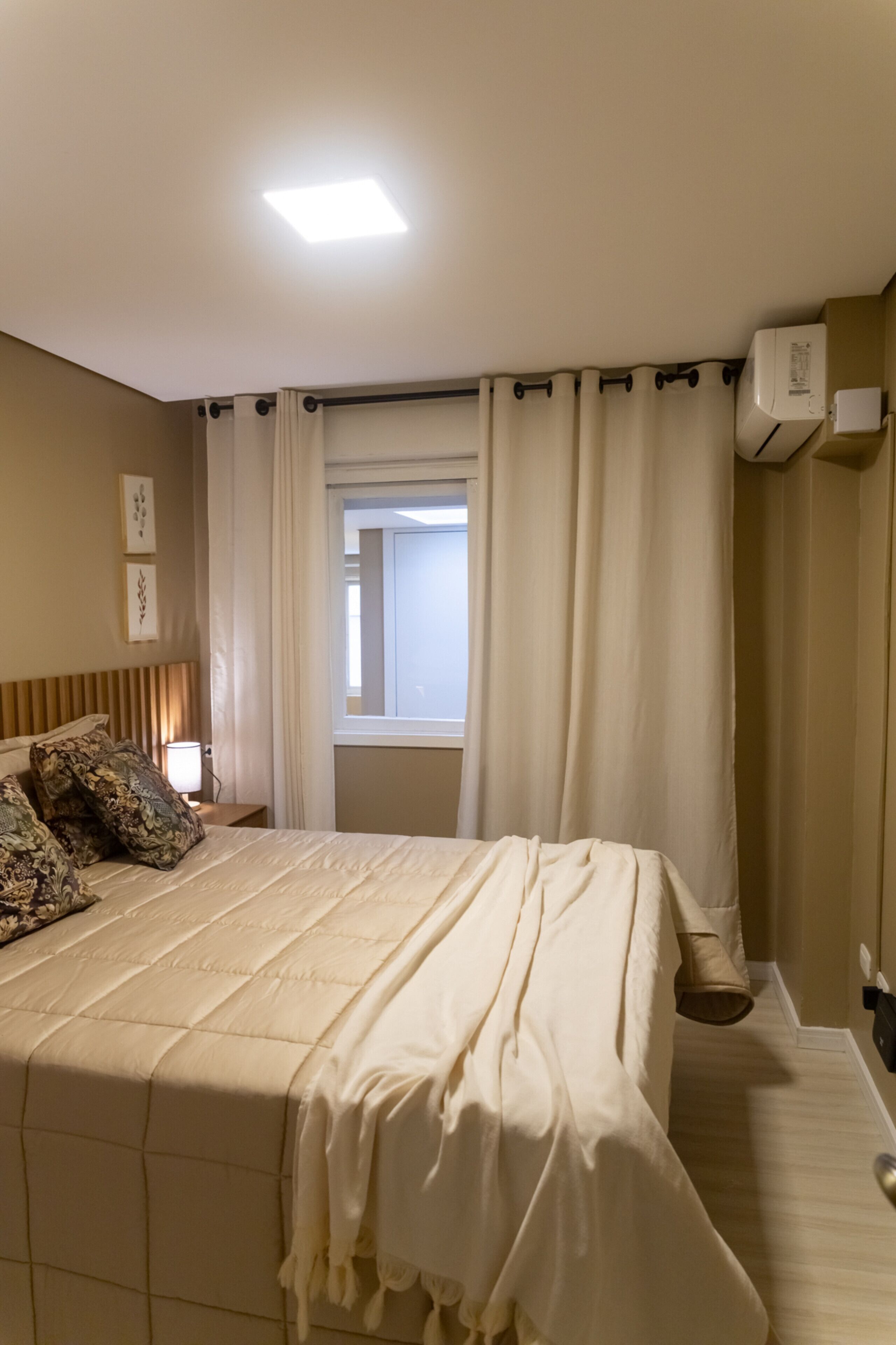 A bedroom with a large bed, a window with curtains, a bedside lamp, and an air conditioning unit.