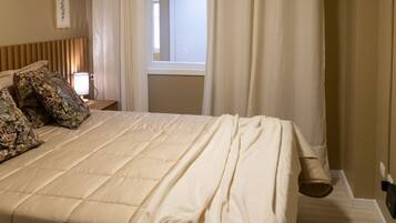A bedroom with a large bed, a window with curtains, a bedside lamp, and an air conditioning unit.