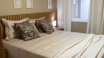 A neatly made bed with a beige comforter and decorative pillows, a bedside table with a lamp, and a window with curtains.