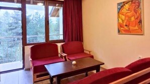 A room with wooden ceiling beams, a large window with red curtains, a painting on the wall, and a seating area with red cushions.
