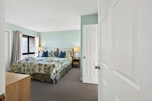 A bedroom with a bed, bedside tables, a nightstand, a door, and a window with curtains. - Poolside Villa Walk To The Beach (Hilton Head Island)