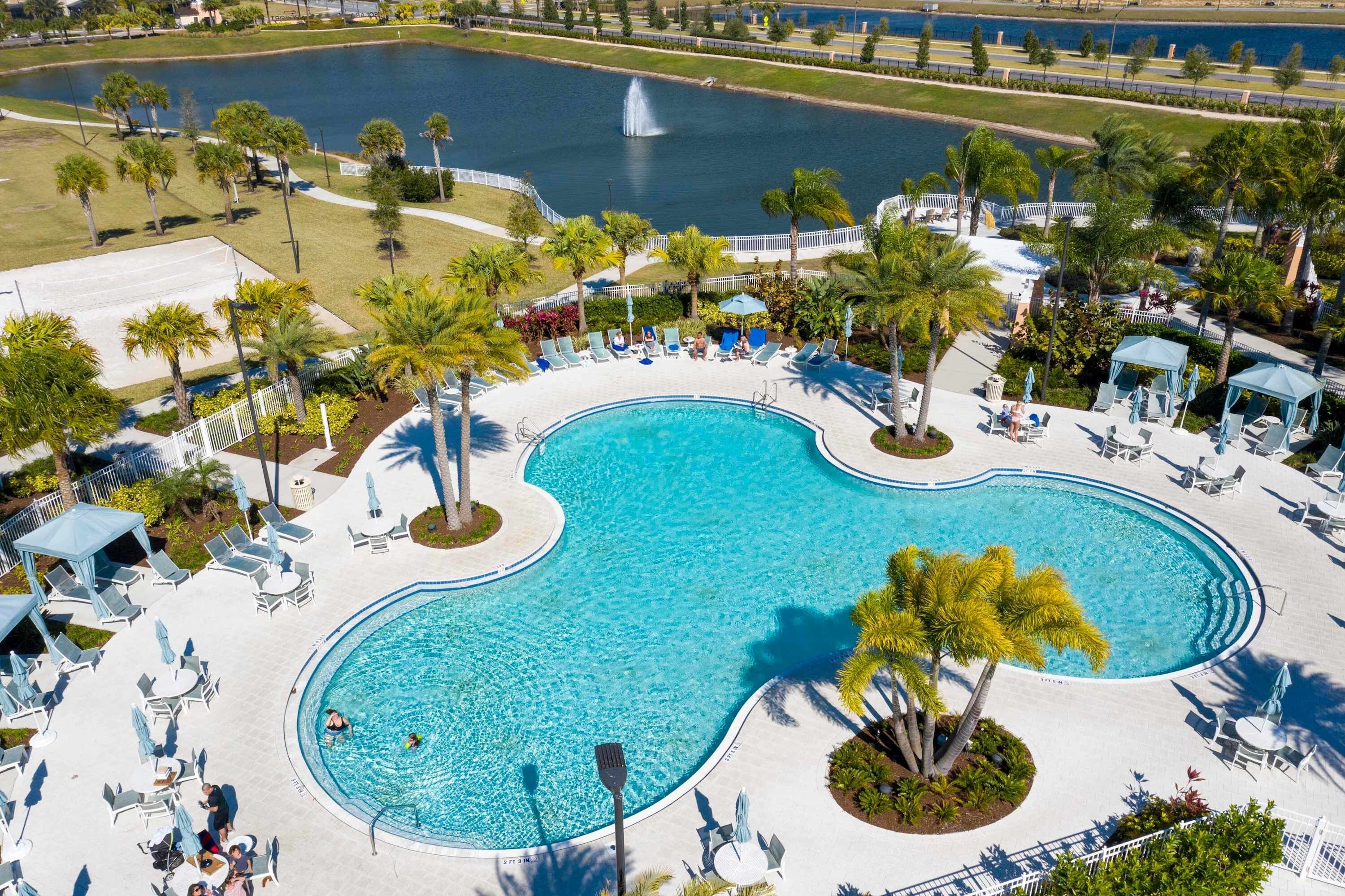 A large, kidney-shaped swimming pool surrounded by lounge chairs and umbrellas, with palm trees and a fountain in the background.