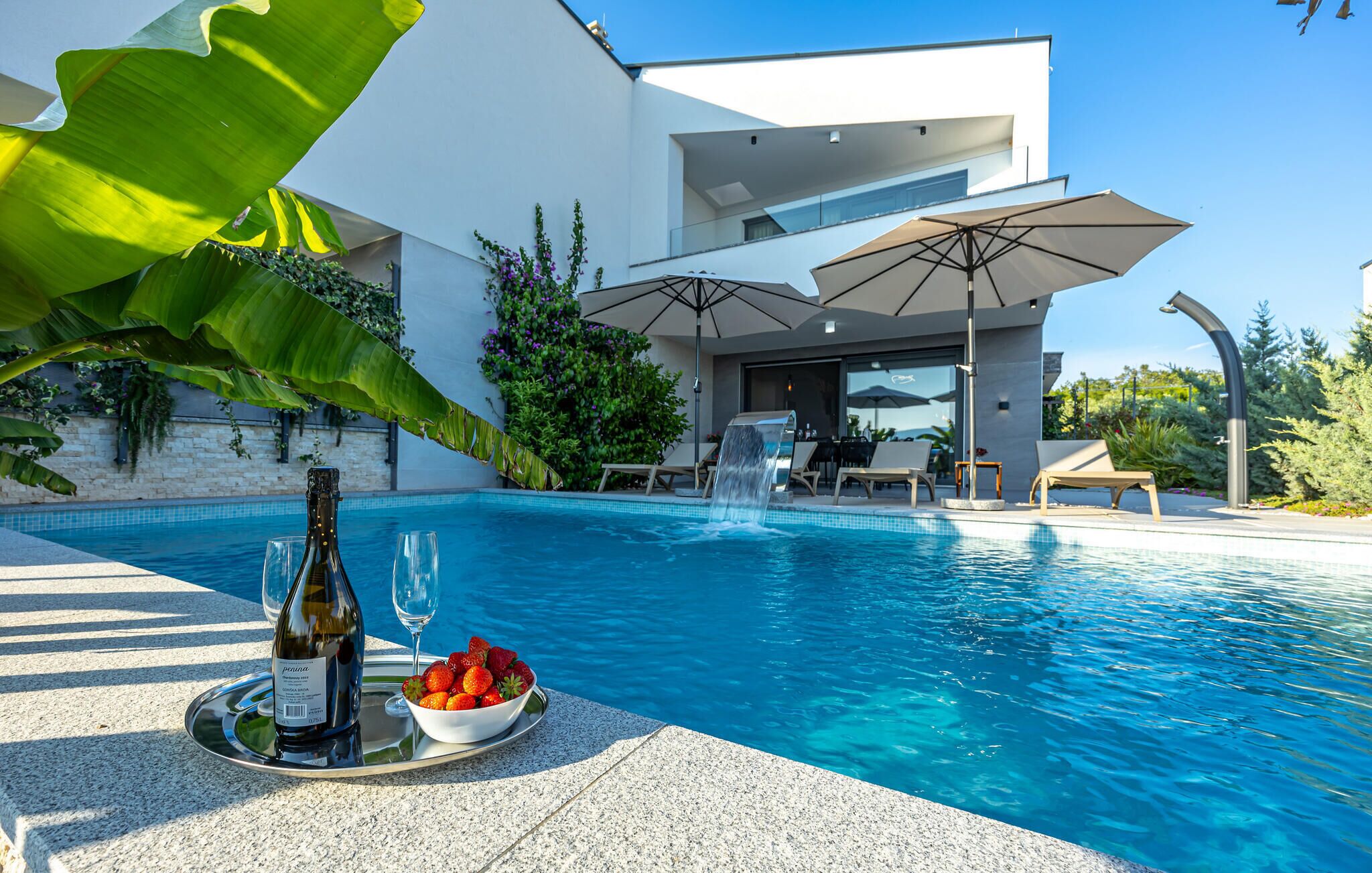 Area piscina moderna con piscina azzurra cristallina, una bottiglia di vino e una ciotola di fragole su un vassoio.