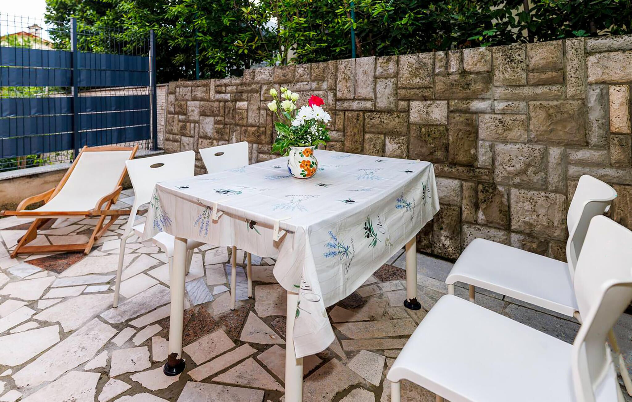 A patio with a stone floor, a table with a floral tablecloth, white chairs, and a potted plant with flowers.
