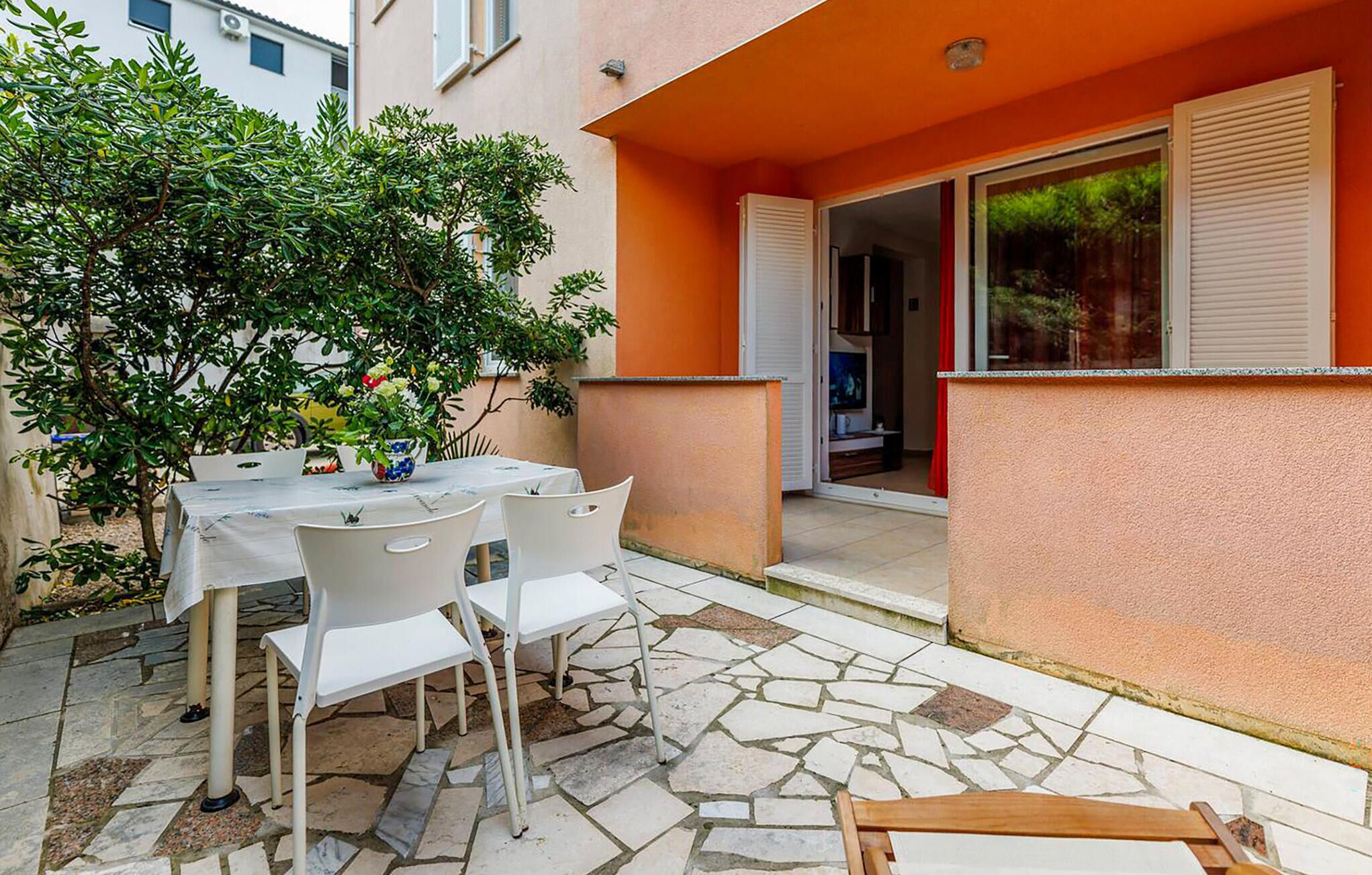 Un patio al aire libre con una mesa y sillas, una planta en maceta y un edificio con paredes anaranjadas y puerta corrediza de vidrio.