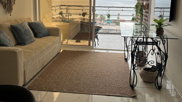 Een woonkamer met een bank, een salontafel, een televisie, een balkon met uitzicht op zee, en een vloerkleed met dessin.