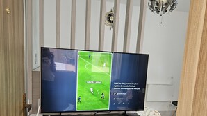 Et fjernsyn på et træstel med en fodboldkamp på skærmen, et ur på væggen og en hængende lampe.
