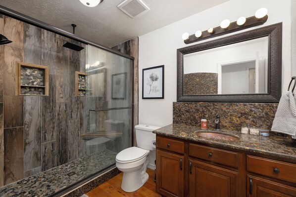 Une salle de bain moderne avec une douche à parois de verre, des toilettes blanches, des meubles en bois et un comptoir en granit.