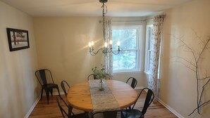 Uma sala de jantar com uma mesa redonda de madeira, cadeiras pretas, um lustre, uma janela com cortinas e um quadro pendurado na parede.