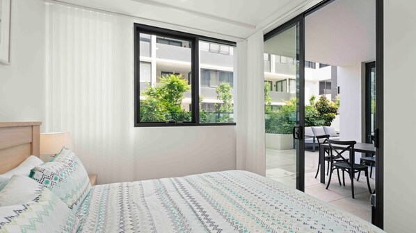 A modern bedroom with a large bed, a window with a view of greenery, and a sliding glass door leading to a balcony with a table and chairs. - Fyve at Allura by Experience Jervis Bay (Huskisson)