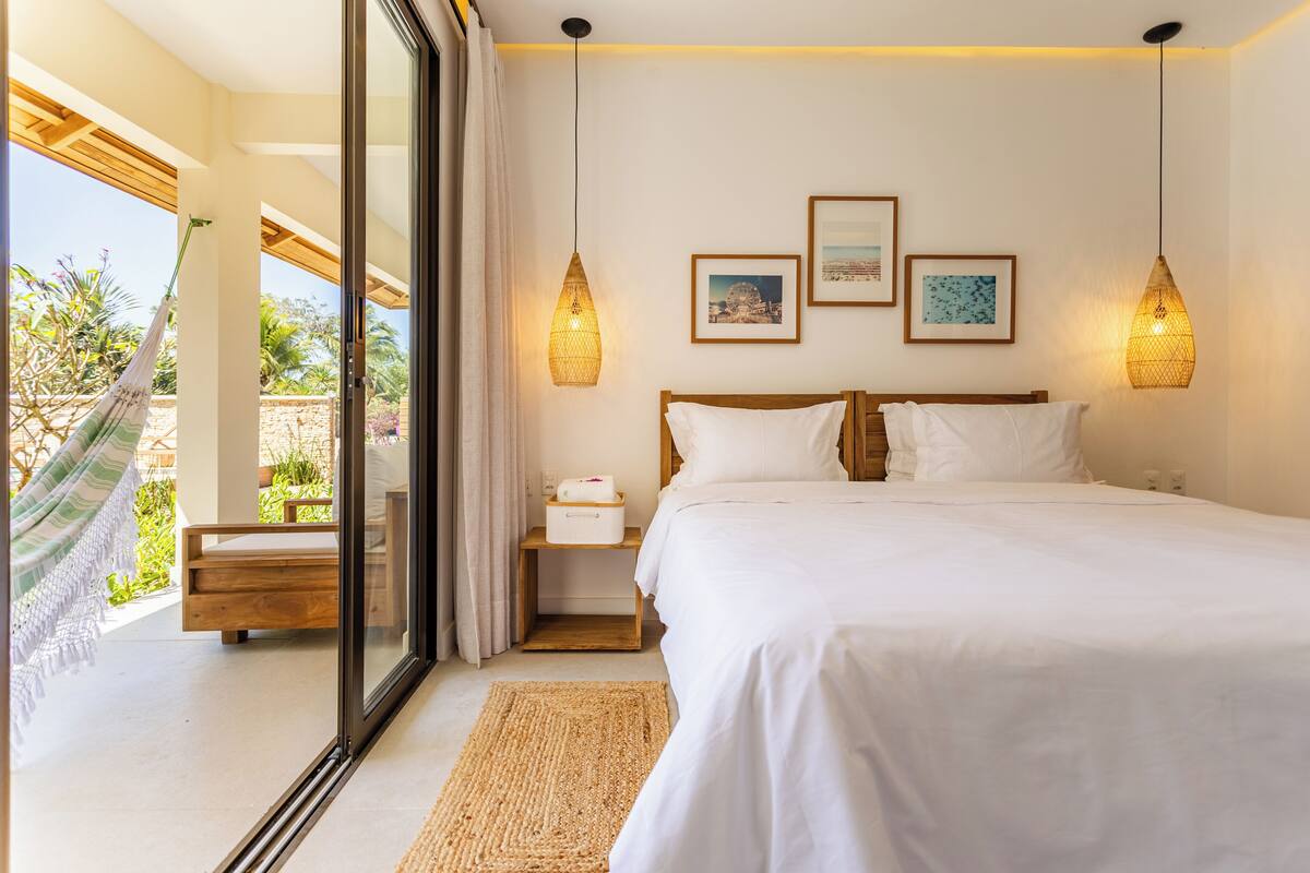 Ein Schlafzimmer mit einem großen Bett, einem Holz-Kopfteil und zwei Wandleuchten. Ein kleiner Nachttisch mit einer Lampe, und ein Blick ins Freie durch eine Glastür.