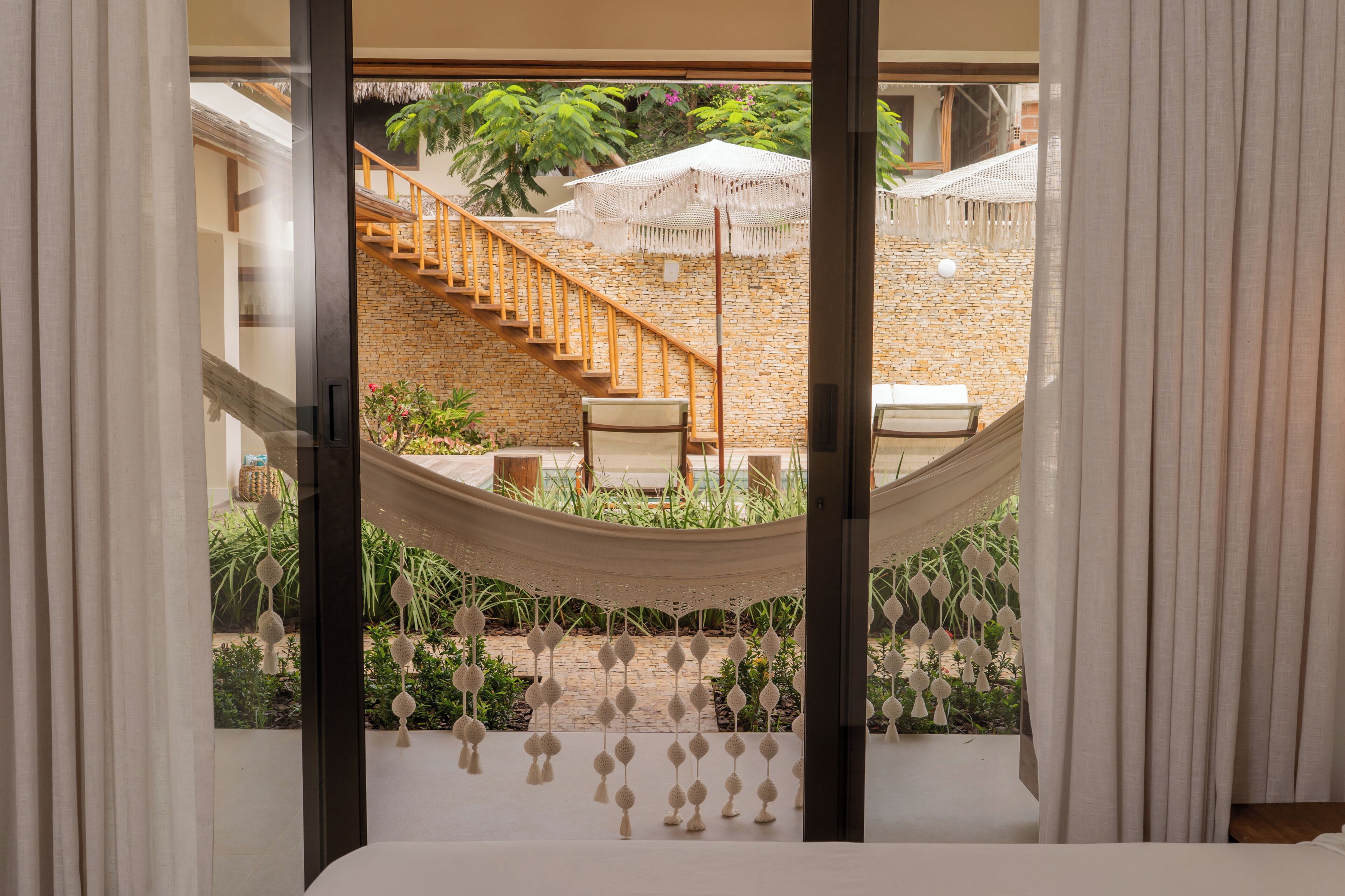 Una hamaca colgada entre dos ventanas, con vista a un muro de ladrillo, una escalera y mobiliario exterior.