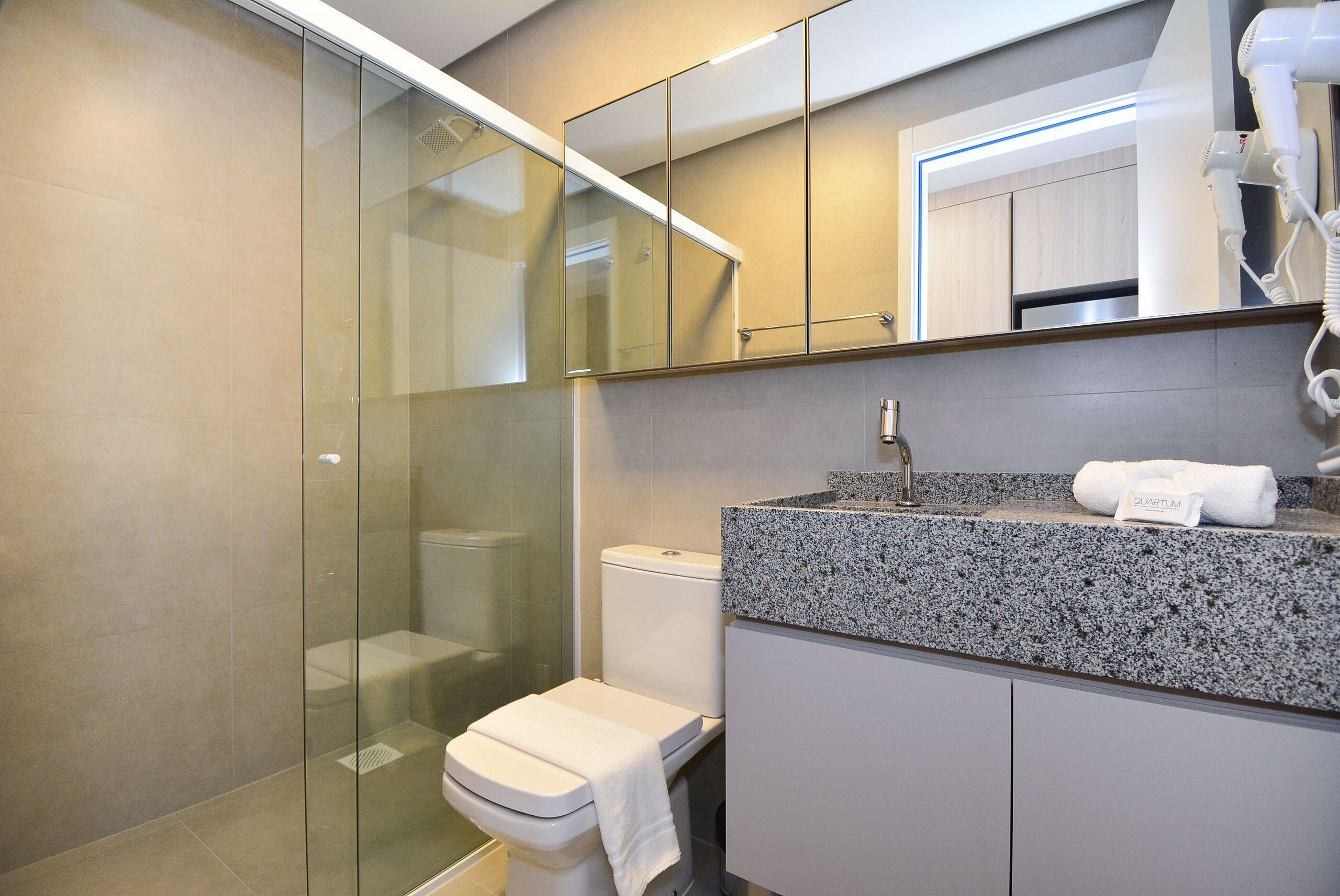A modern bathroom with a glass shower enclosure, a toilet, a granite countertop with a sink, and a hairdryer on the counter.