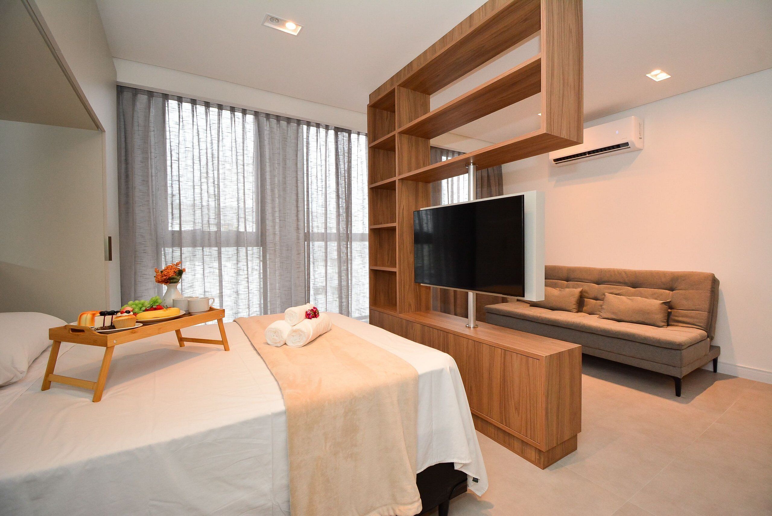 A modern hotel room with a bed, a wooden side table with a tray, a sofa, a TV, and an air conditioning unit.