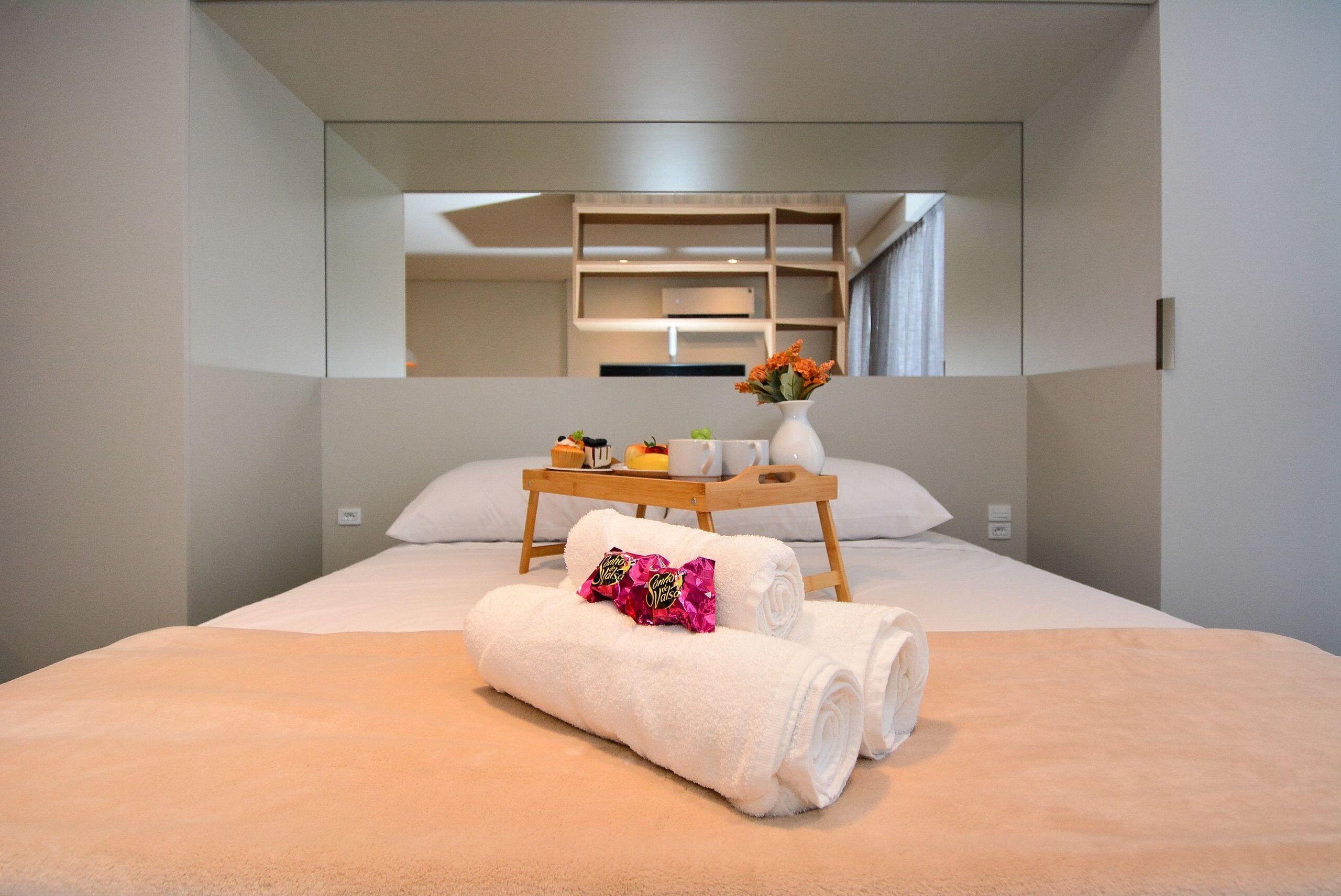 Una habitación de hotel moderna con una cama, una mesita de noche con un jarrón de flores y un espejo que refleja el interior de la habitación.
