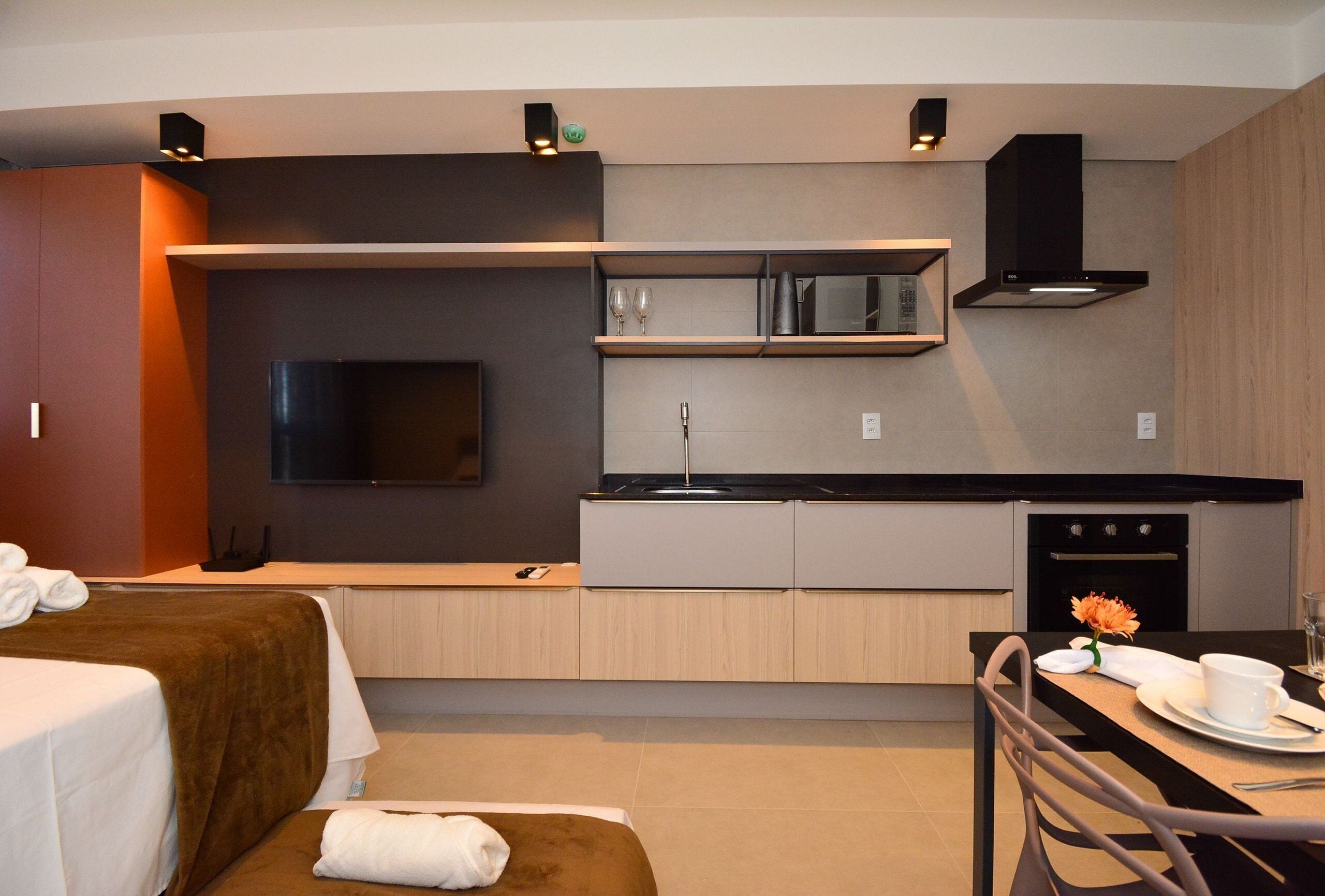 A modern hotel room with a kitchenette, a bed with a brown headboard, a dining area with a table and chairs, and a flat-screen TV mounted on the wall.