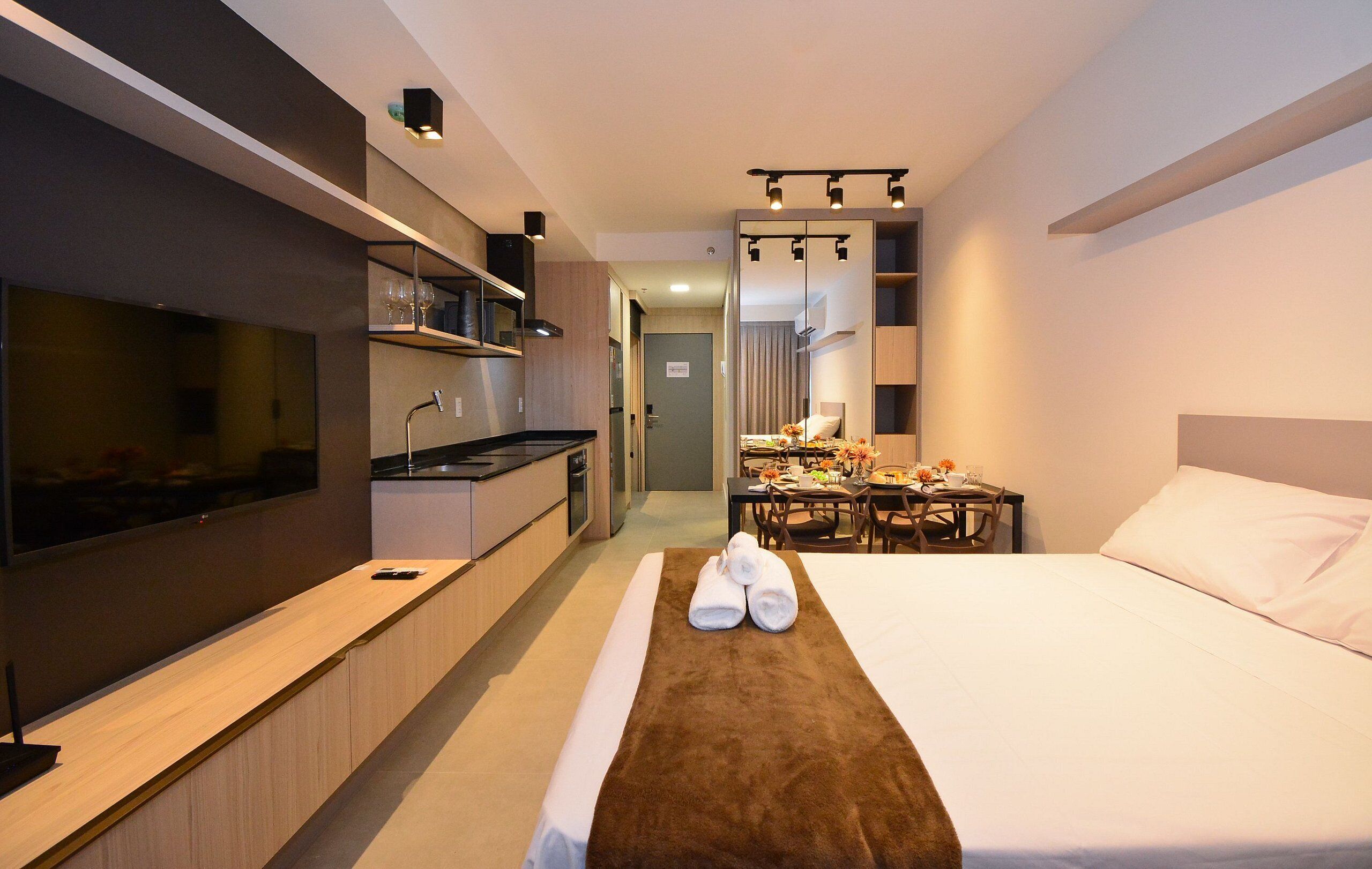 A modern hotel room with a bed, a TV, a kitchenette, and a dining area.