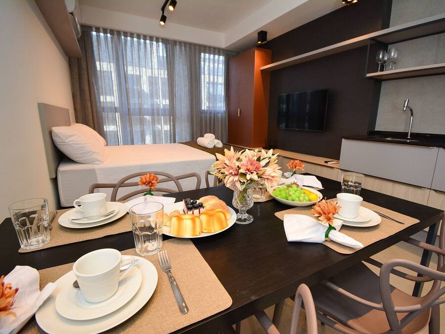 Habitación de hotel moderna con cama, una mesa de comedor con desayuno servido, zona de cocina y televisor montado en la pared.