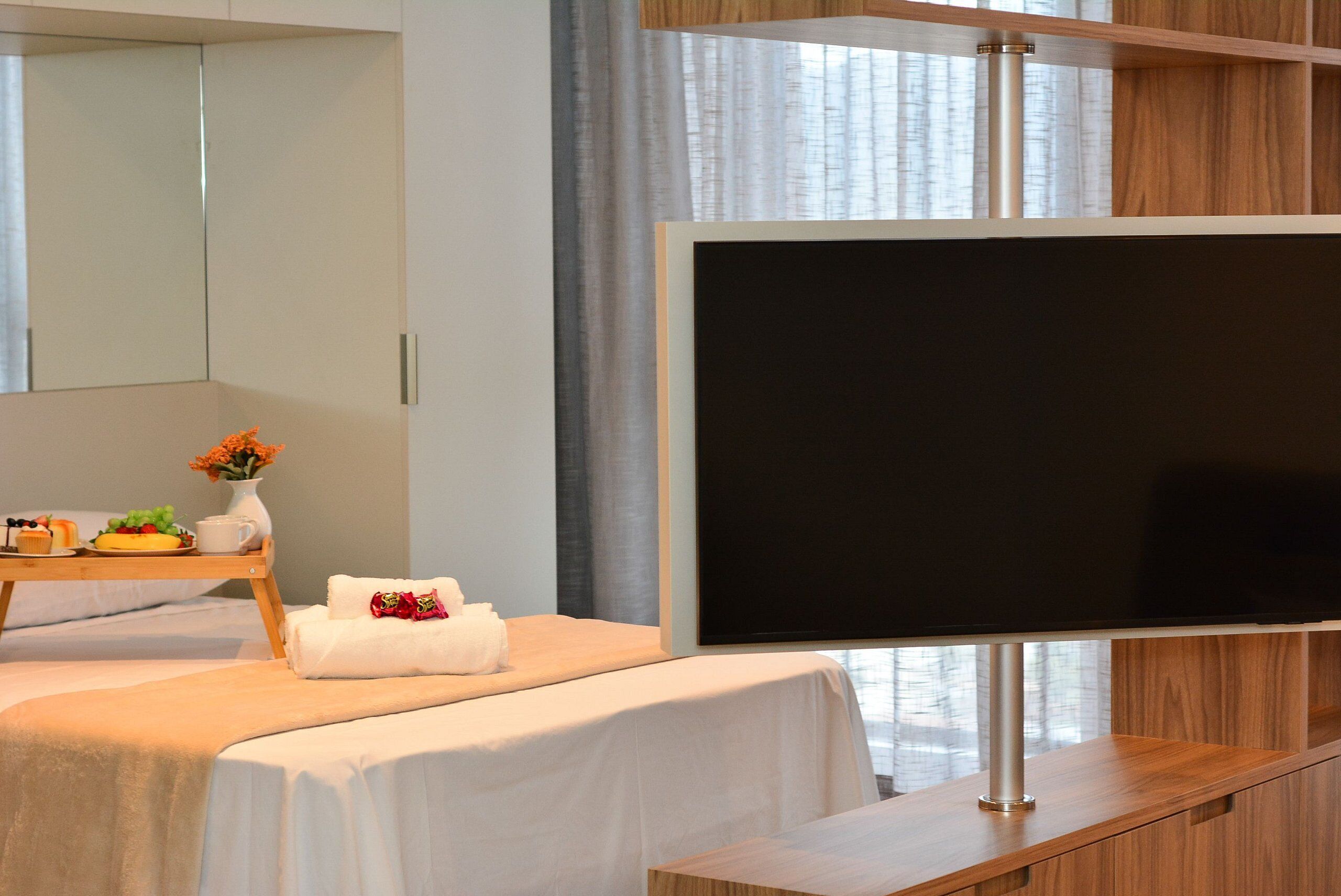 Ett modernt hotellrum med en säng, en platt-TV, ett litet sidobord i trä med en vas med blommor och en stor spegel.