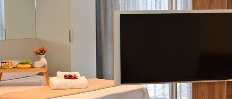 Ett modernt hotellrum med en säng, en platt-TV, ett litet sidobord i trä med en vas med blommor och en stor spegel.