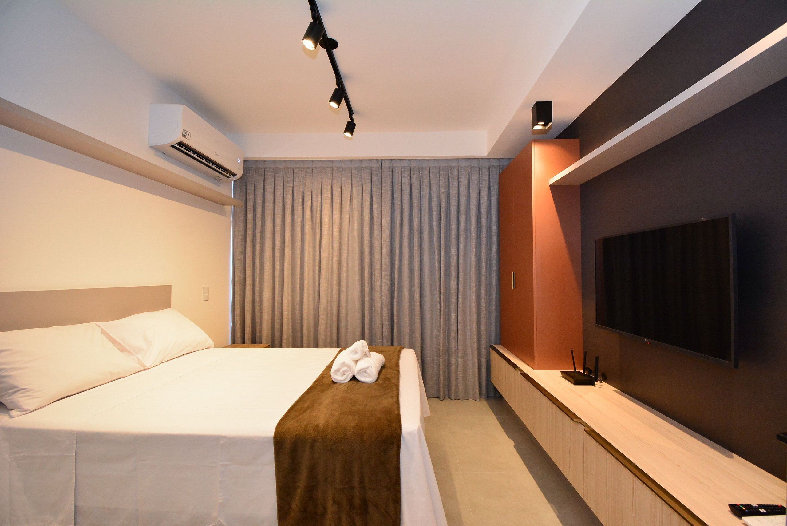A hotel room with a bed, a flat-screen TV, an air conditioning unit, and a bedside table with a remote control.