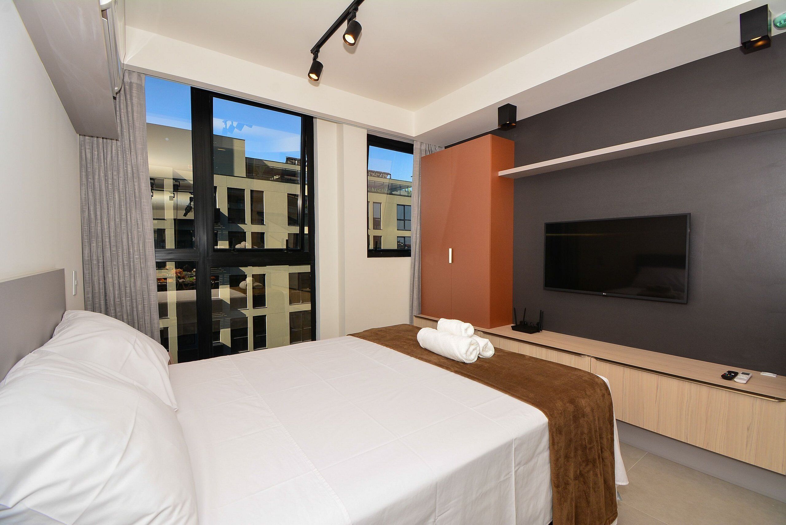 A hotel room with a bed, a TV, a sliding glass door, and a view of buildings outside.