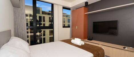 A hotel room with a bed, a TV, a sliding glass door, and a view of buildings outside.