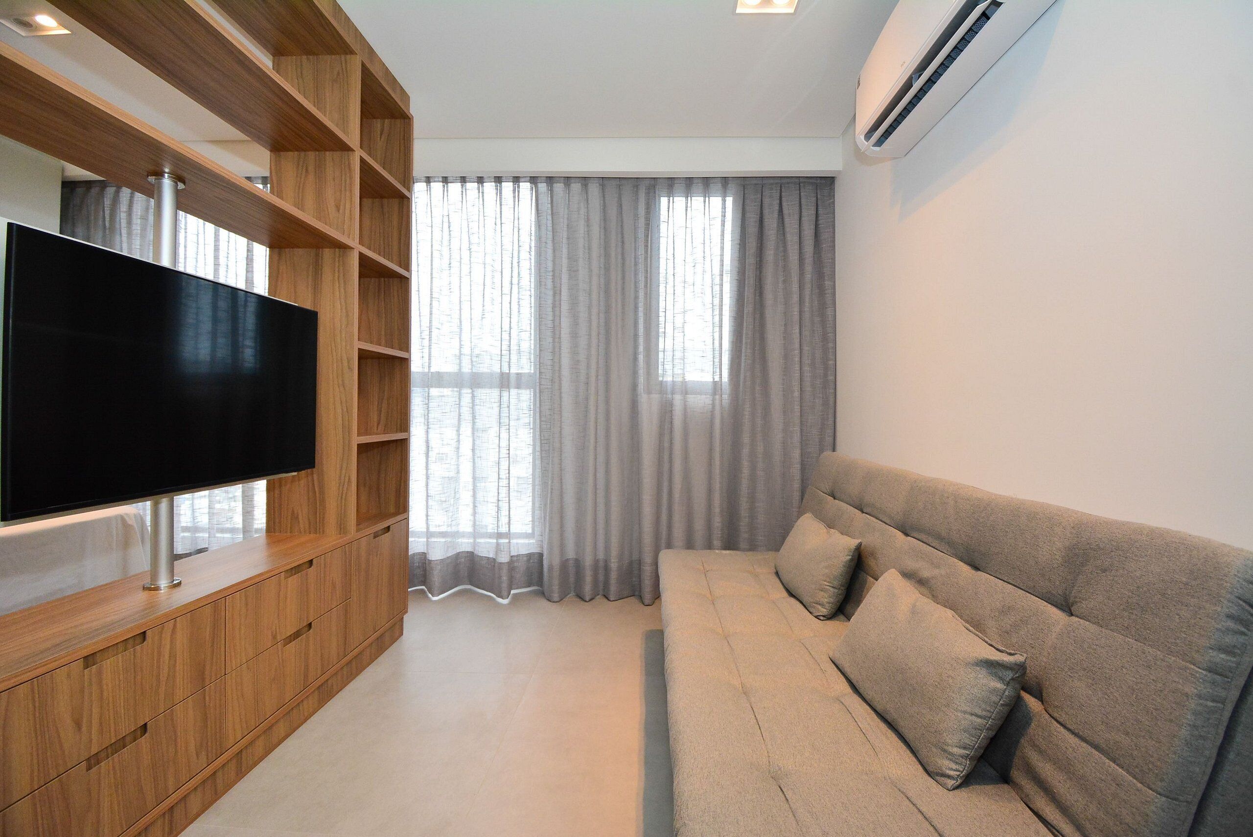 A modern living room with a flat-screen TV, wooden shelving, a grey sofa, and a large window with curtains.