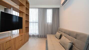 A modern living room with a flat-screen TV, wooden shelving, a grey sofa, and a large window with curtains.