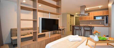 A modern hotel room with a bed, a TV, a kitchenette, and a seating area.