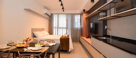 A modern hotel room with a dining table set for breakfast, a bed with a white headboard, a kitchen area with a microwave, and a large window with sheer curtains.