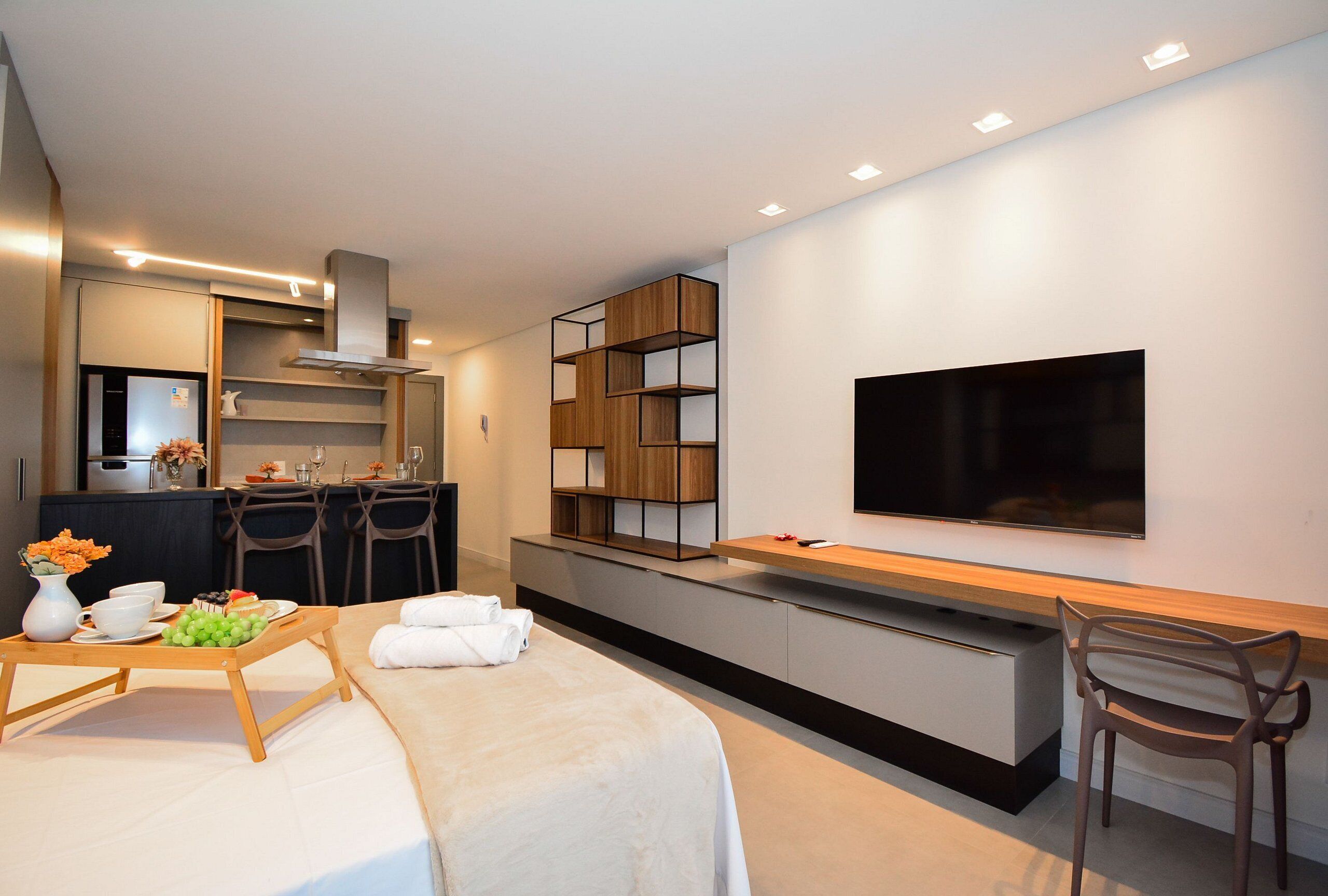 A modern hotel room with a bed, a dining area with a table set for two, a kitchen area, and a flat-screen TV mounted on the wall.