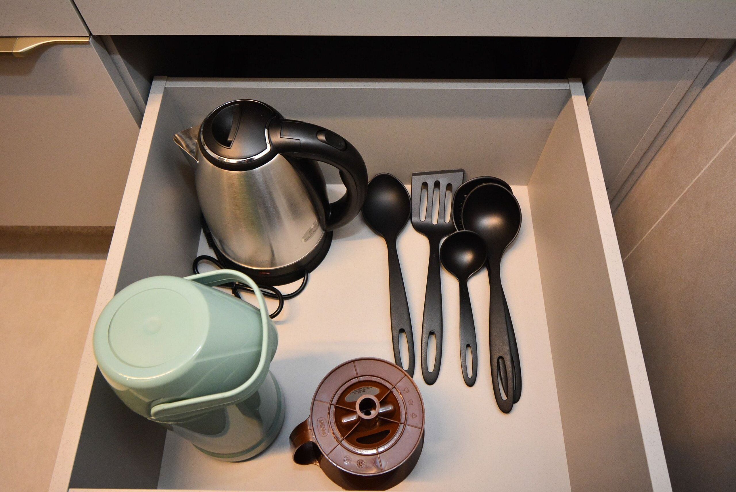 Un cajón con una tetera de acero inoxidable, una tetera verde, una cafetera marrón y utensilios de cocina negros.