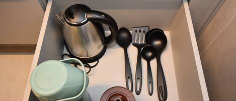 A drawer containing a stainless steel kettle, a green teapot, a brown coffee pot, and black kitchen utensils.