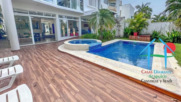 Ett modernt hus med en swimmingpool, ett trädäck och glasdörrar som leder in till ett rum med möbler.
