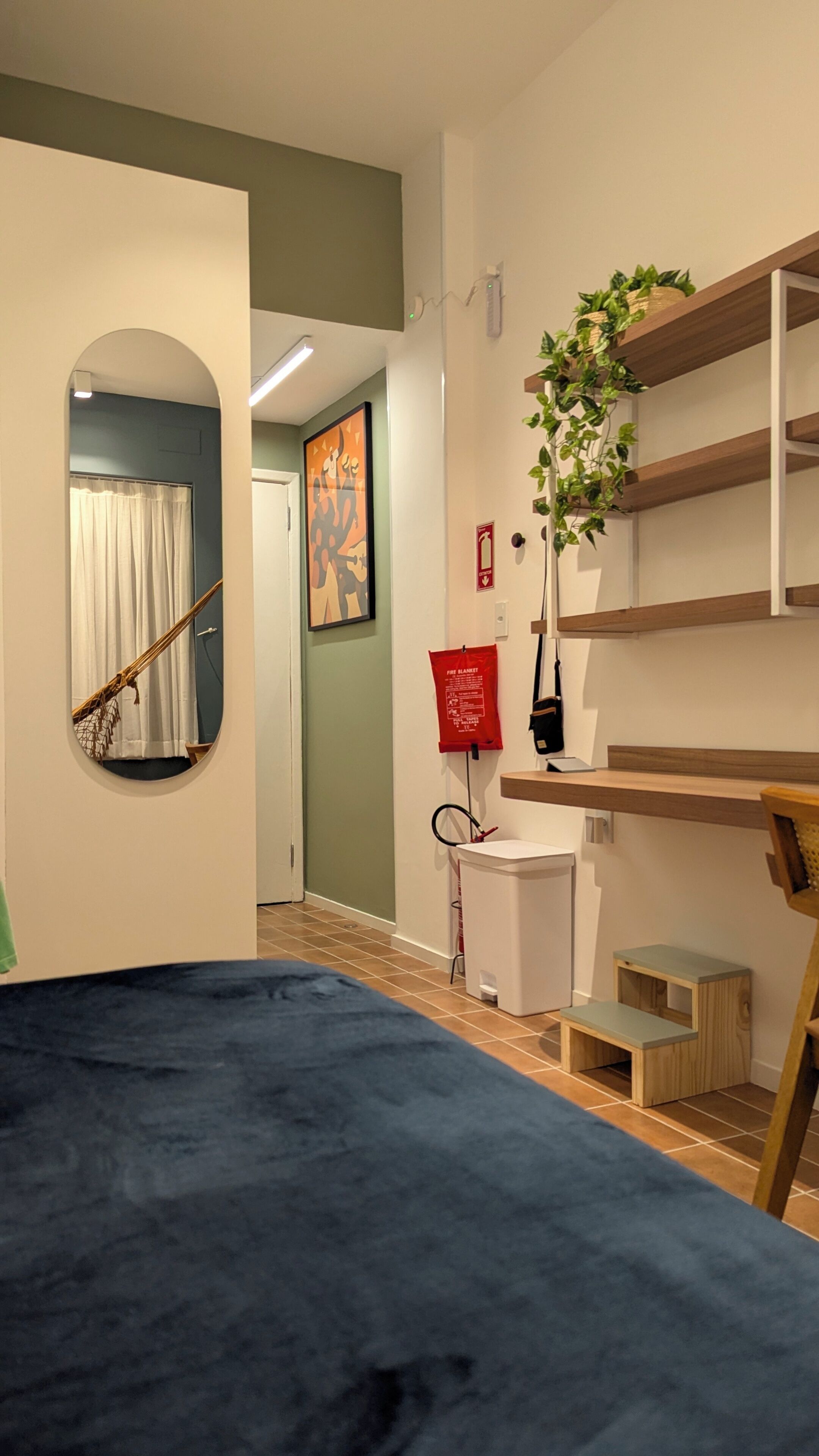 A room with a blue bed, a wooden shelf unit, a small wooden stool, a fire blanket, and a plant.