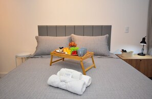 Een netjes opgemaakt bed met een grijs hoofdeinde, een nachtkastje met een lamp en een dienblad met fruit en een fles op het bed.