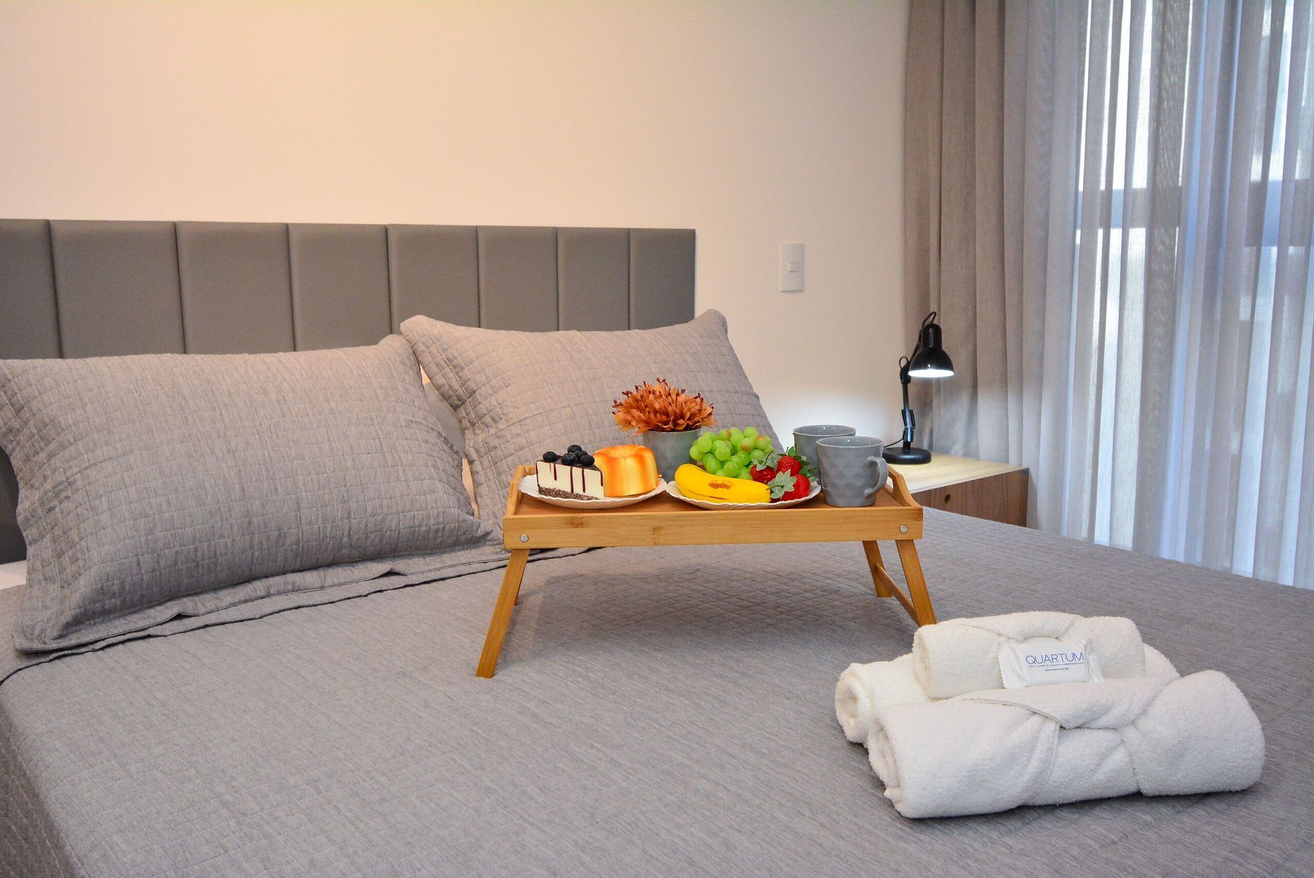 A neatly made bed with a tray of fruit and a cake, a folded towel, and a bedside lamp.