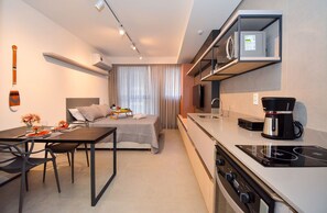 Een compacte hotelkamer met een bed, een zithoek met een tafel en stoelen, een kitchenette met een fornuis en koffiezetapparaat, en een televisie op een plank.