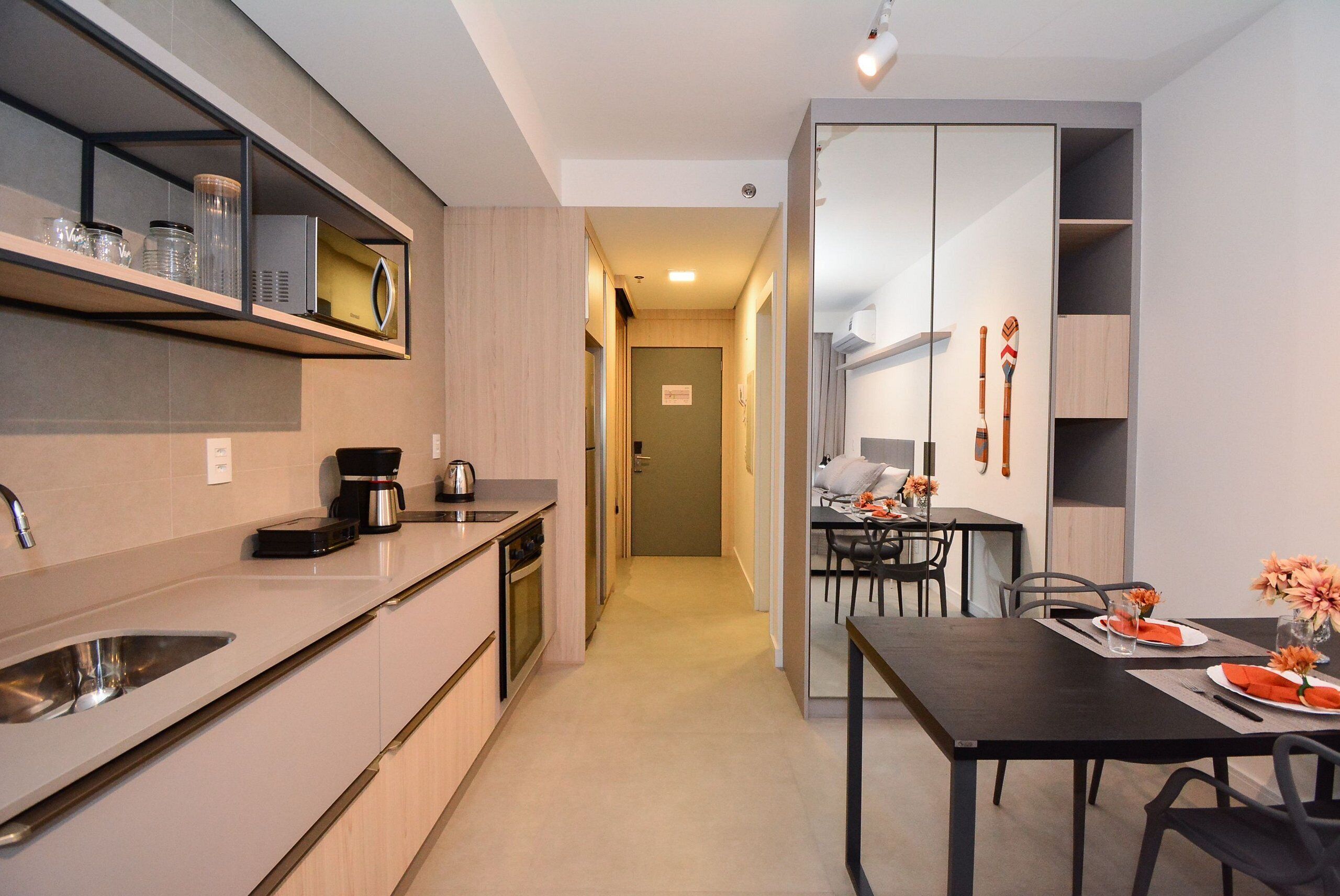 Una cocina moderna con horno integrado, cafetera y lavamanos. Hay un comedor con una mesa preparada para dos personas, y un espejo en la pared.