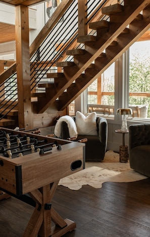 Eine Holztreppe, ein Kicker, eine Couch und eine Lampe in einem Raum mit großen Fenstern.