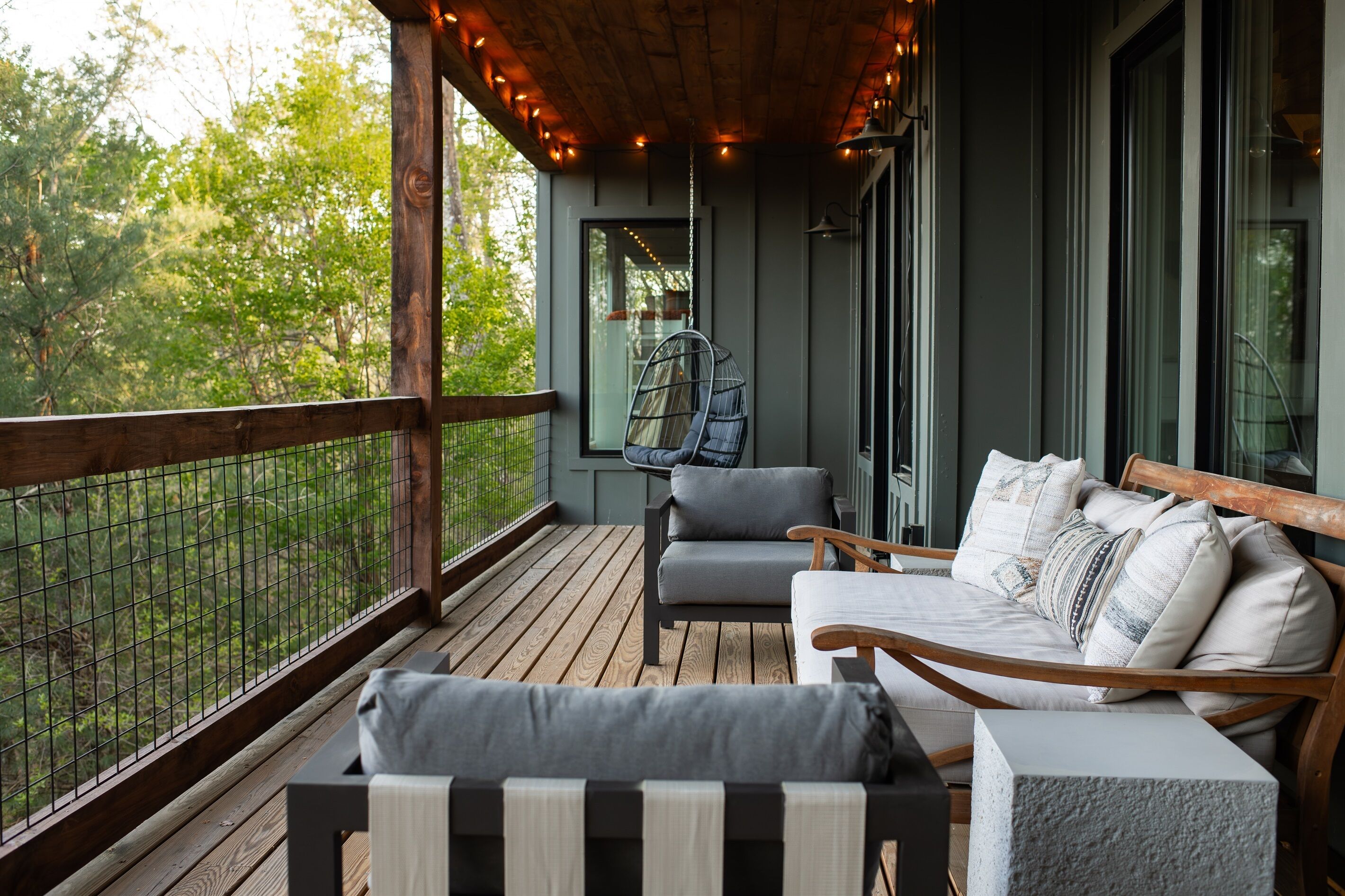 A wooden deck with a sofa and chairs, a hanging chair, and a view of greenery.