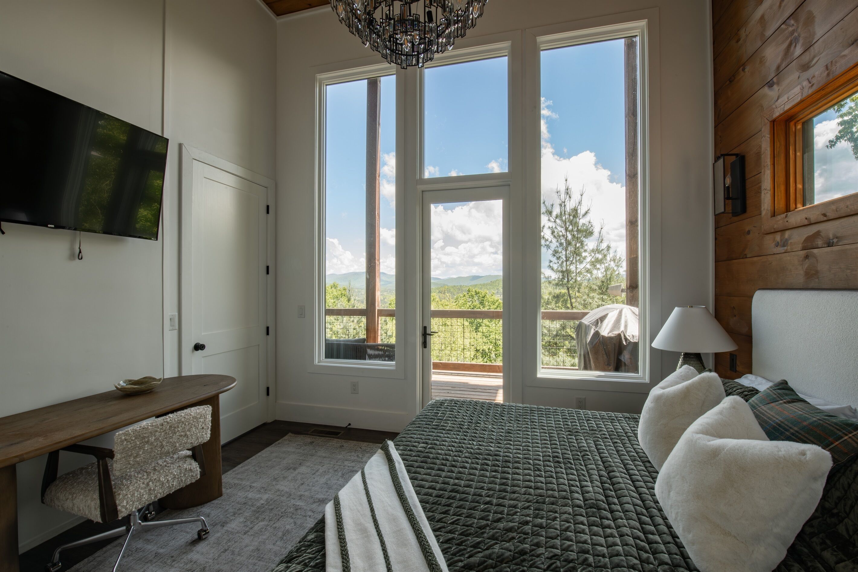 A bedroom with a bed, a desk, a TV, and large windows with a view.