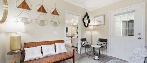 A modern living room with a brick-patterned sofa, a fluffy grey sofa, a small round table, and a chair. There are three pendant lights on the wall, a large mirror, and a door with a window.